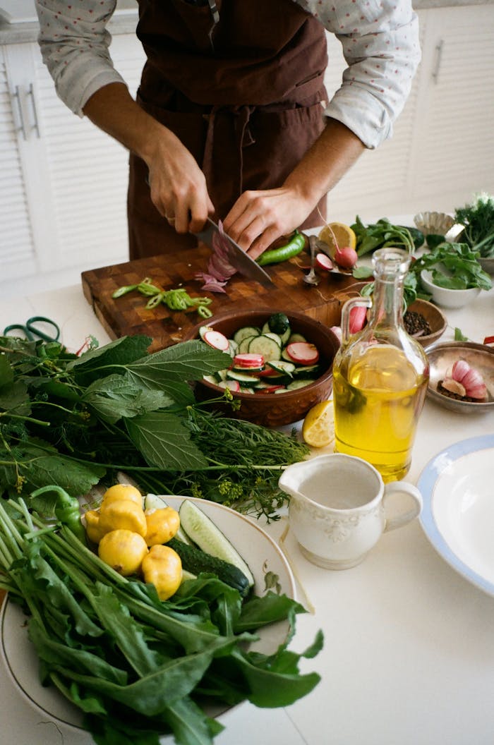 gallery-01 Adult preparing salad with fresh vegetables, knife, and olive oil in kitchen setting.